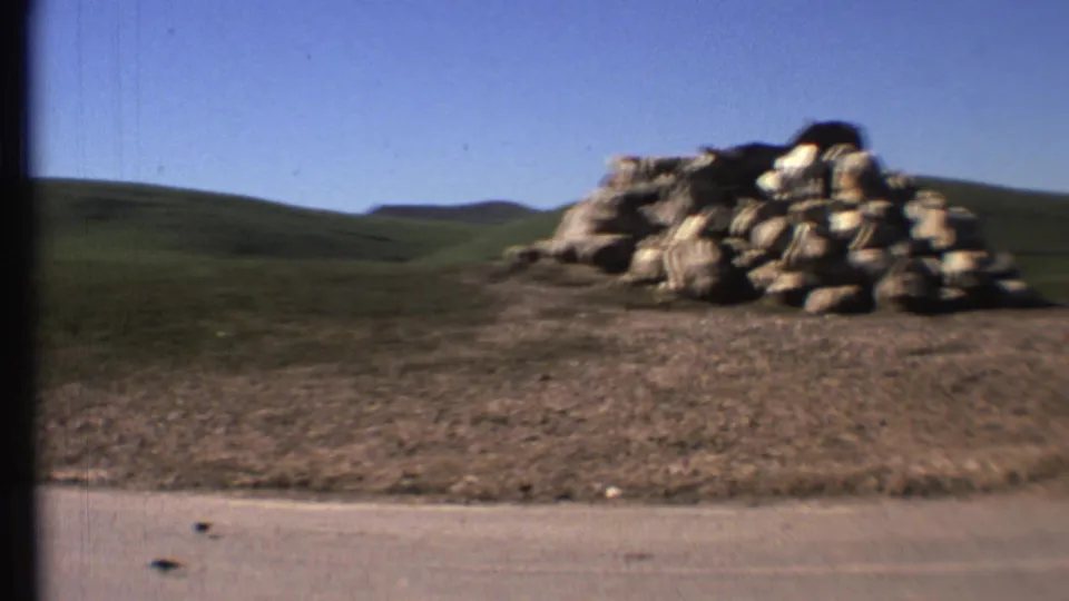 The final frame holds on the paved road, grassy hillside, blue sky, and a large rock pile after the donkey has moved out of view.