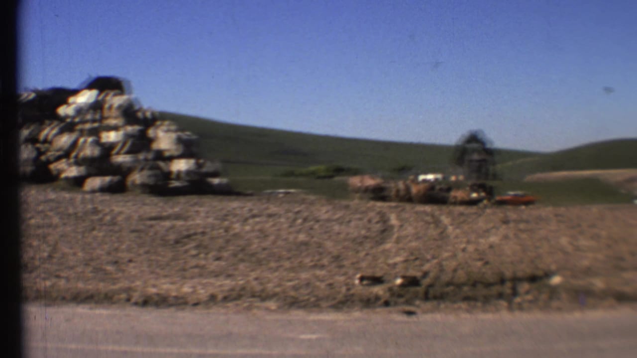 Roadside rock pile with open hills, cattle, a small structure, and vehicles in the distance.