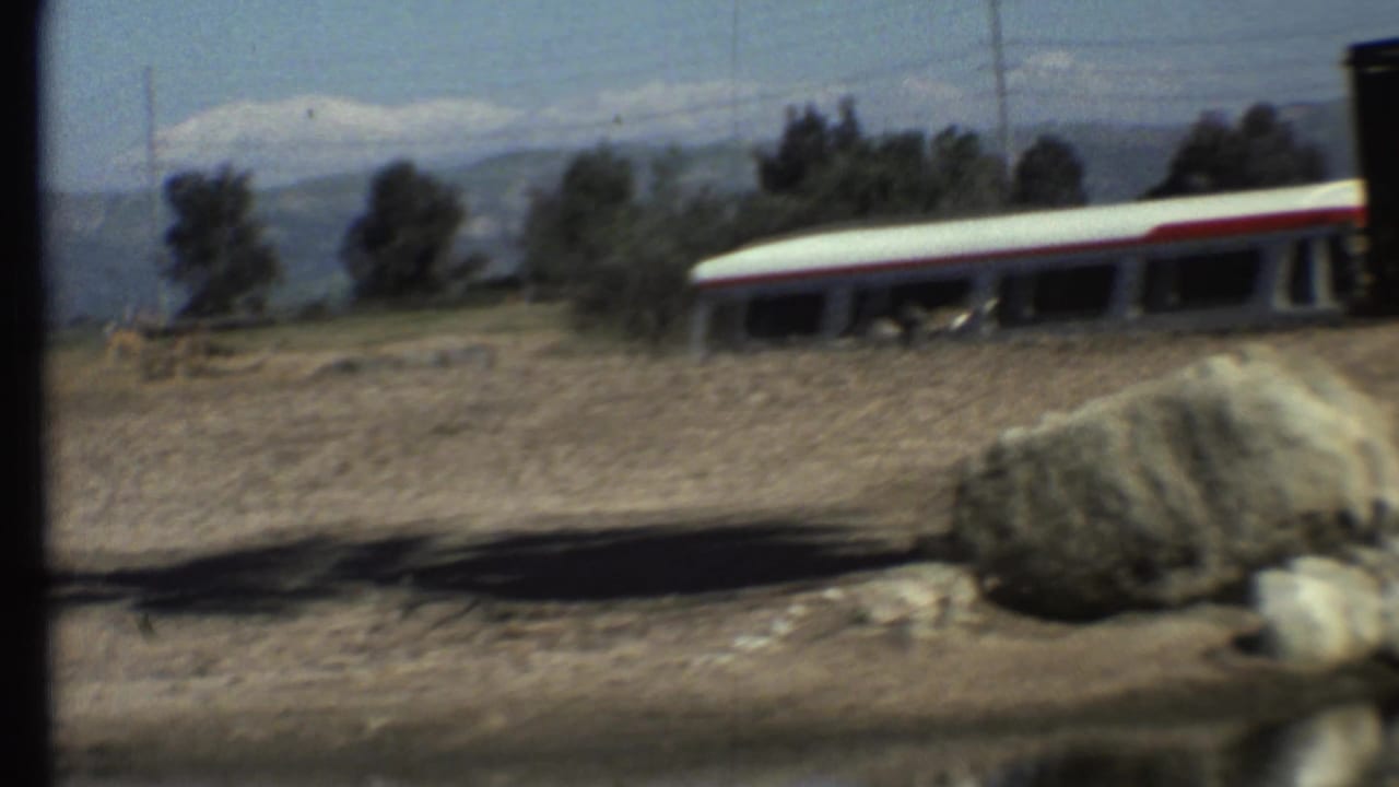 1972 Grand Canyon-area frame of a red-striped bus crossing an arid roadside scene with rocks, trees, and mountains.
