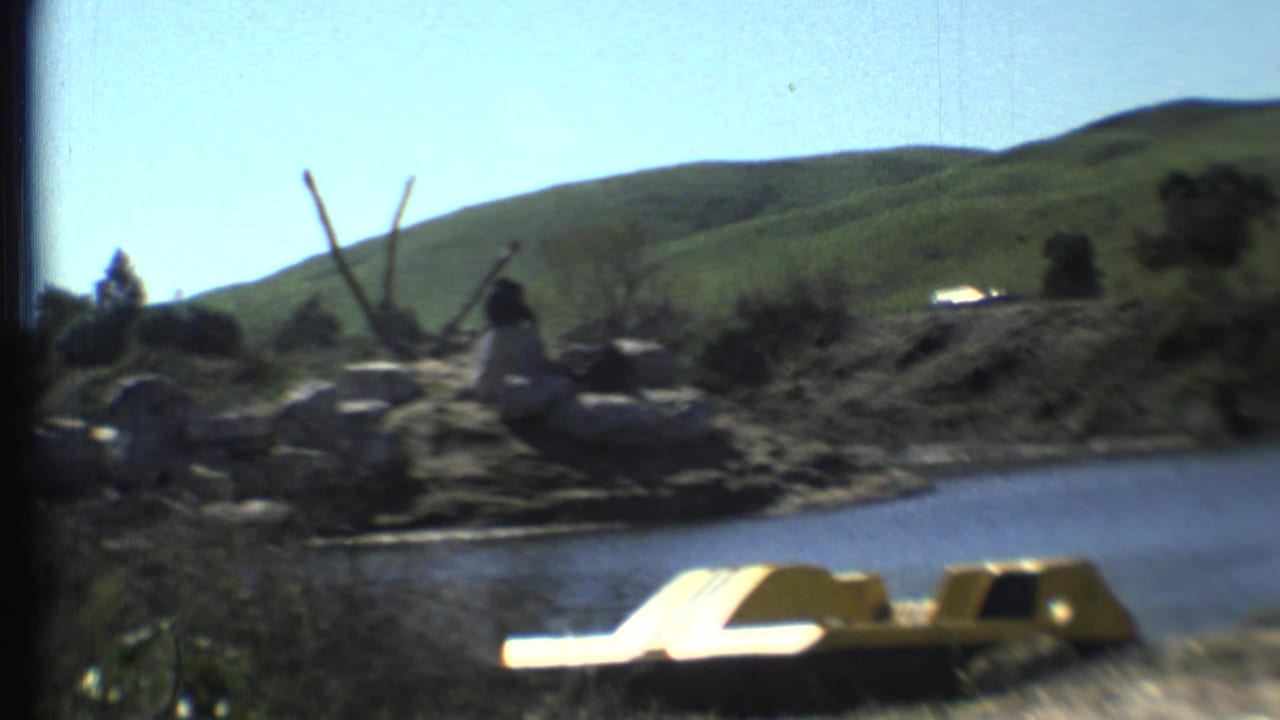 Tiger lies near a rocky water edge with a yellow paddle boat and green hills behind it.