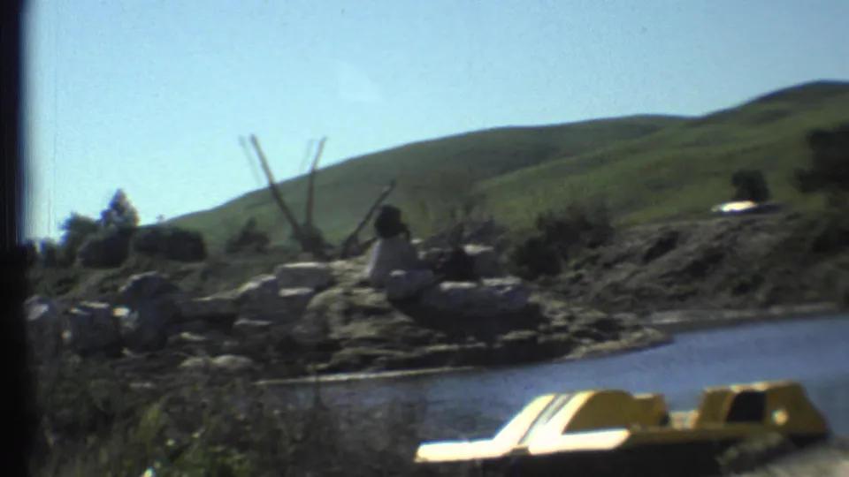 Vintage 8mm frame shows a tiger on a rocky bank above water, with a yellow paddle boat and green hills in the background.