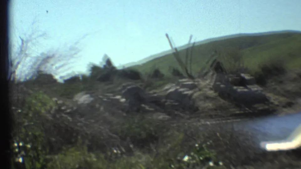 The handheld 8mm view drifts across the rocky animal enclosure area, showing water, brush, hills, and part of a yellow boat.