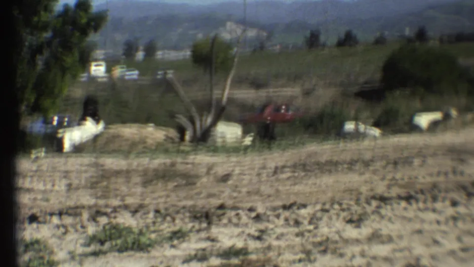 1972 8mm travel footage shows a dirt foreground, distant parked cars, small roadside structures, trees, and hazy hills.