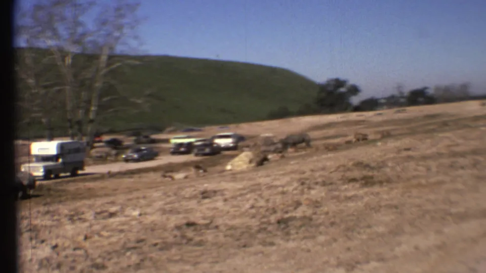 1972 Grand Canyon-area home-movie frame showing a dry roadside field with classic cars, a white camper van, grazing animals, trees, and a green hill.