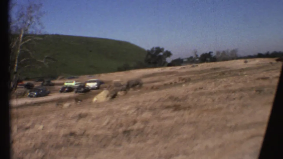 1972 Arizona travel footage frame with cars clustered near a dirt road as large animals graze across a dry field below a green hillside.