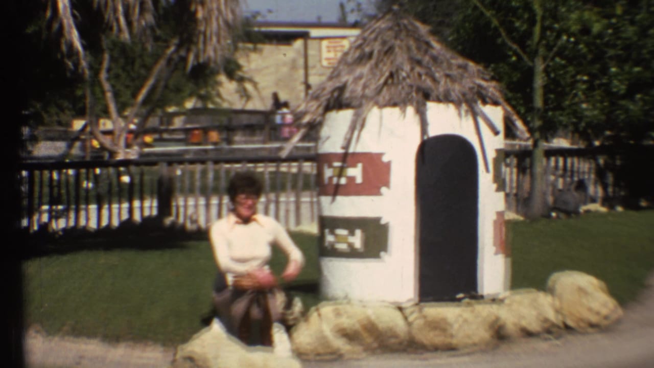 Woman sitting on rocks beside a small thatched-roof structure in a grassy outdoor area.