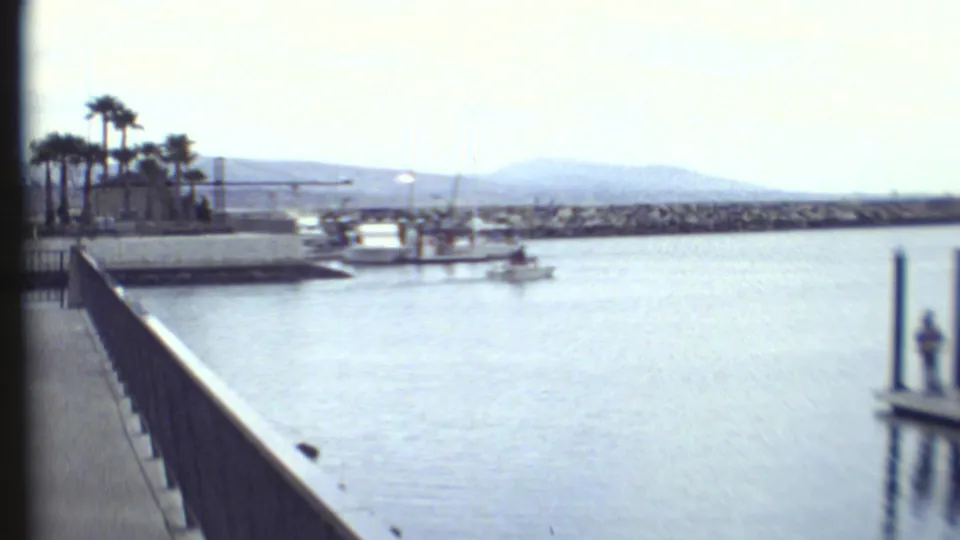 1972 8mm home-movie frame cuts to a waterfront with railing, boats, palm trees, distant mountains, and a figure on a dock.
