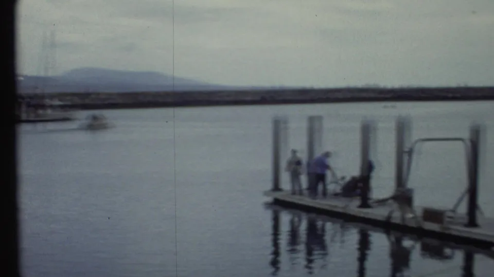 1972 8mm frame showing people on a floating marina dock with pilings, calm harbor water, and hills along the horizon.