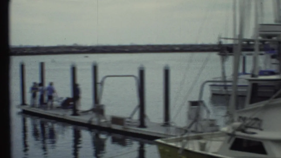 1972 8mm marina frame with people on a floating dock, vertical pilings, nearby boats, rigging, and a low shoreline beyond the water.