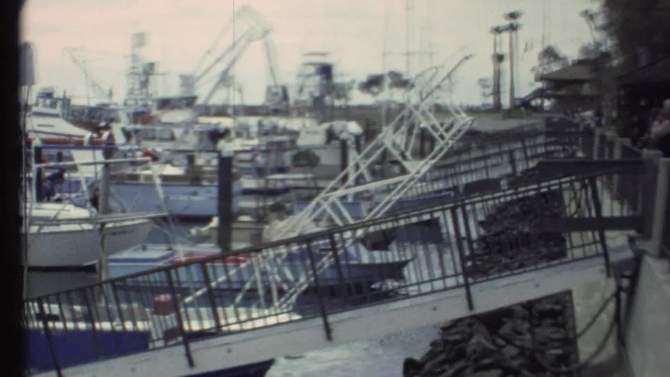 1972 8mm harbor shot with foreground railings and rocks, crowded boats, cranes, and light poles under hazy sky.