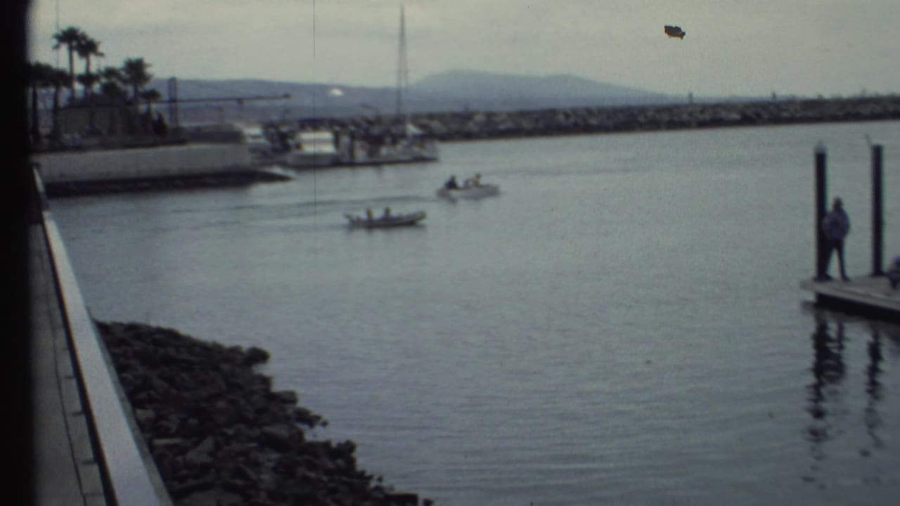 1972 8mm frame of two small motorboats with people aboard moving across harbor water near a rocky breakwater and dock posts.