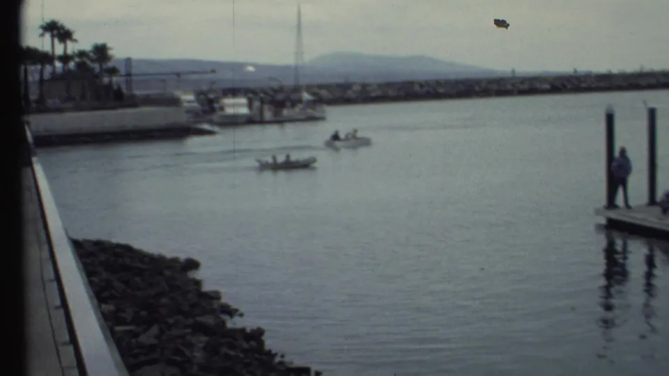 1972 8mm frame of two small motorboats with people aboard moving across harbor water near a rocky breakwater and dock posts.