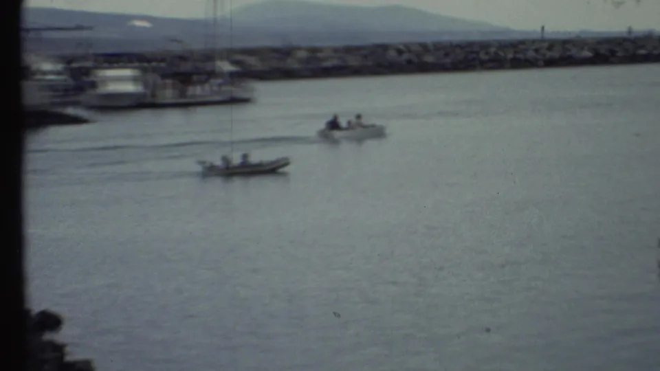 1972 home-movie frame showing two small boats carrying people across calm harbor water near a larger docked vessel.