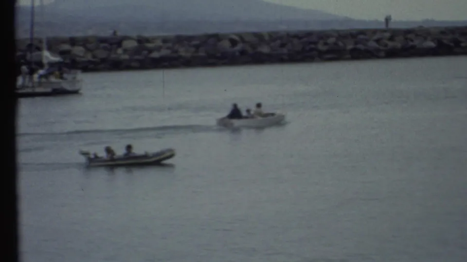1972 8mm frame showing two small motorboats with people aboard crossing harbor water before a rocky breakwater and distant hills.