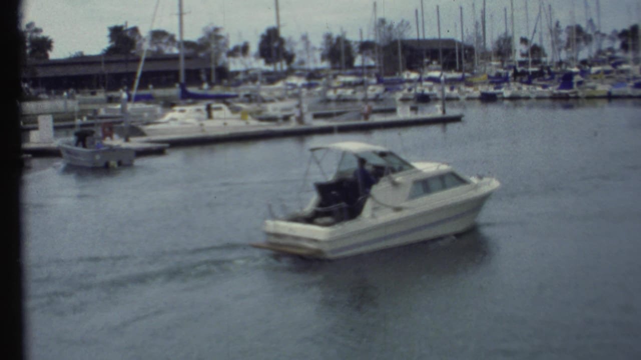 Small cabin motorboat moving away from a marina with docks and sailboats behind it.