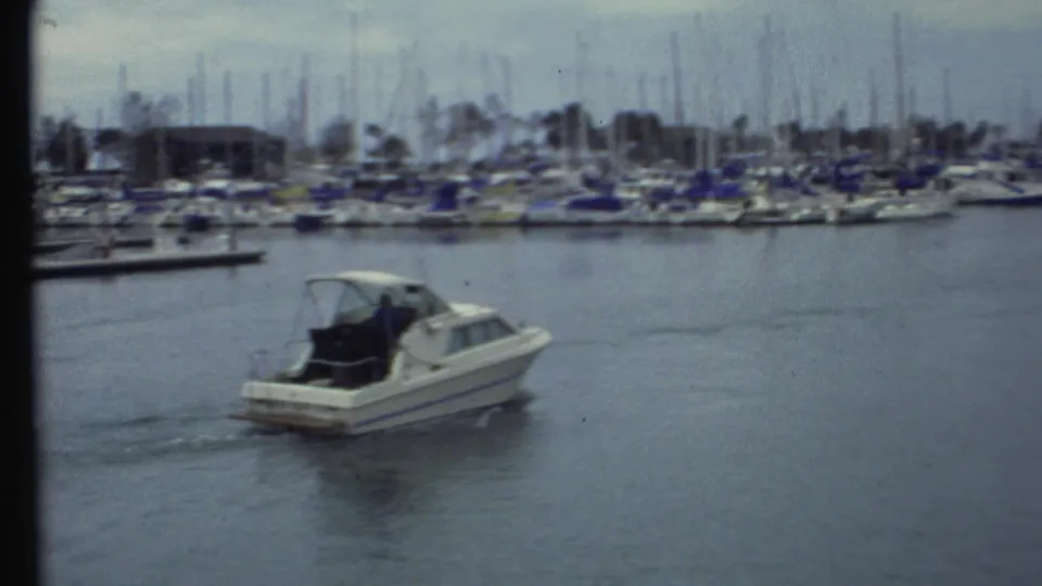 Soft 8mm frame of the motorboat passing through marina water, with moored sailboats and docks across the background.