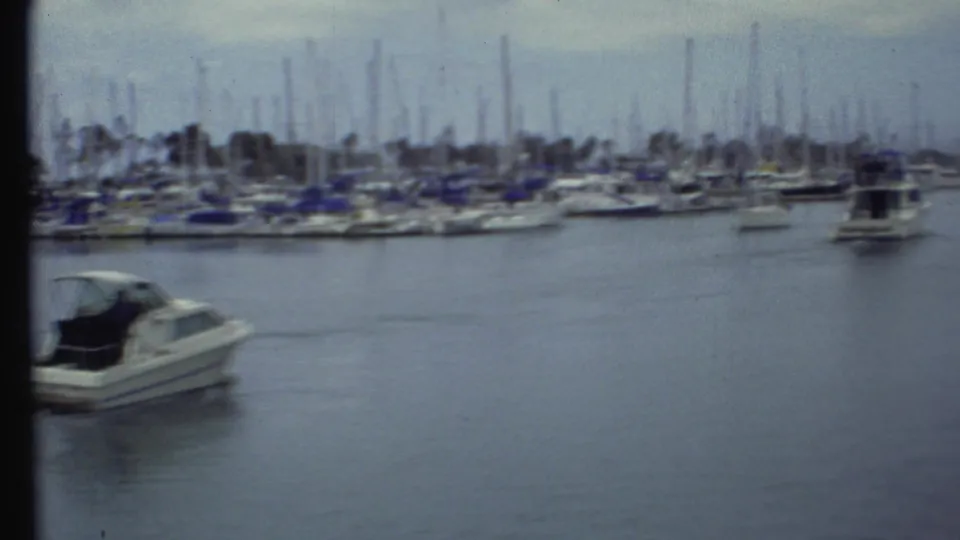 1972 8mm frame with the motorboat exiting left, open water in the foreground, and a crowded marina under gray sky.