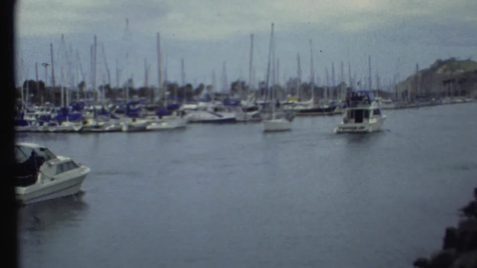 1972 marina footage showing a white motorboat moving through open water in front of clustered docked sailboats.