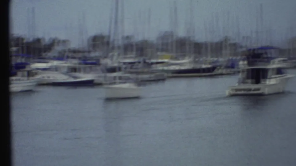 Soft, handheld 1972 8mm frame of a marina with moored sailboats and a white motorboat near the right edge.
