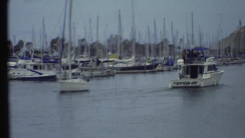 1972 8mm home-movie frame of a marina with docked sailboats, a white motorboat, calm water, and a low hillside in the background.