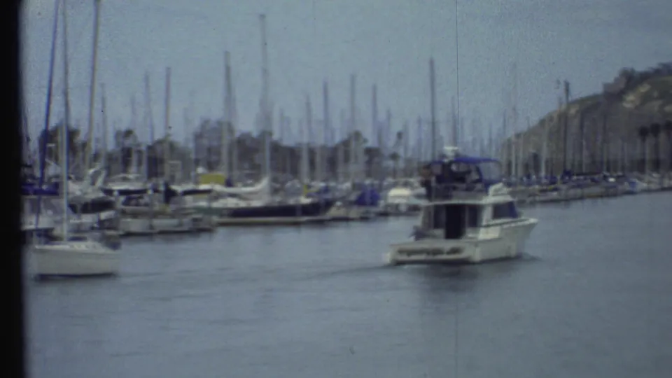 Vintage 8mm frame showing a white motorboat with a blue canopy traveling through a busy marina, with sailboats and hillside behind it.