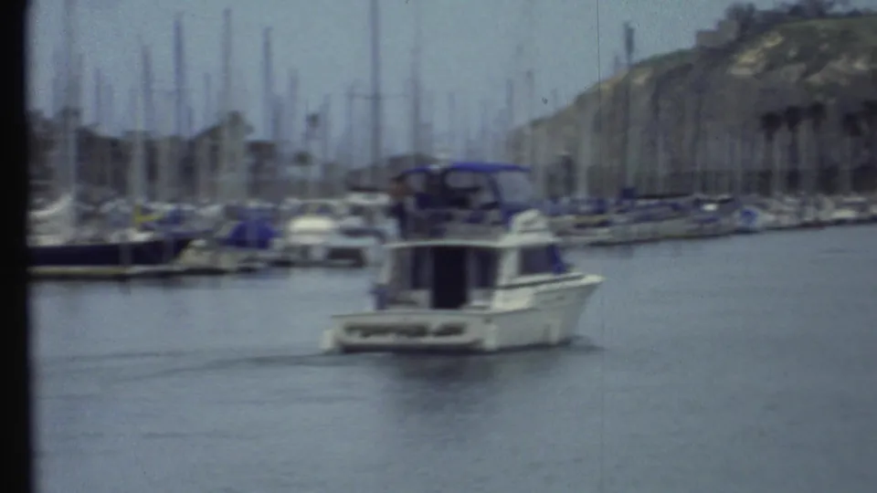 Soft, grainy 1972 home-movie frame of a motorboat crossing calm marina water with blurred sailboats and a hillside beyond.