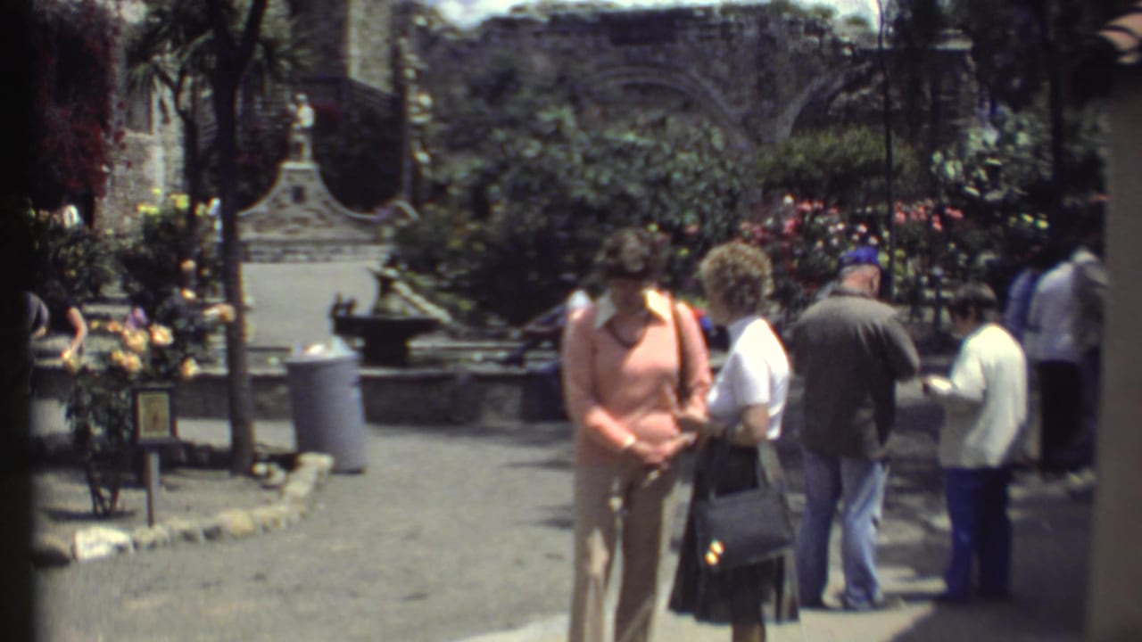 Tourists stand in a sunny landscaped courtyard with flowers, trees, benches, and stone buildings.