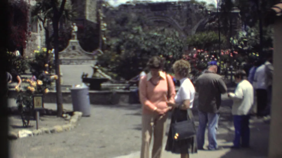 1972 Grand Canyon home-movie frame showing visitors in a planted courtyard with trees, flowers, benches, and stone architecture.