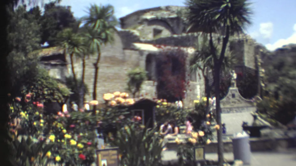 1972 Grand Canyon home-movie frame showing a landscaped courtyard, seated visitors, flower beds, trees, and stone architecture.