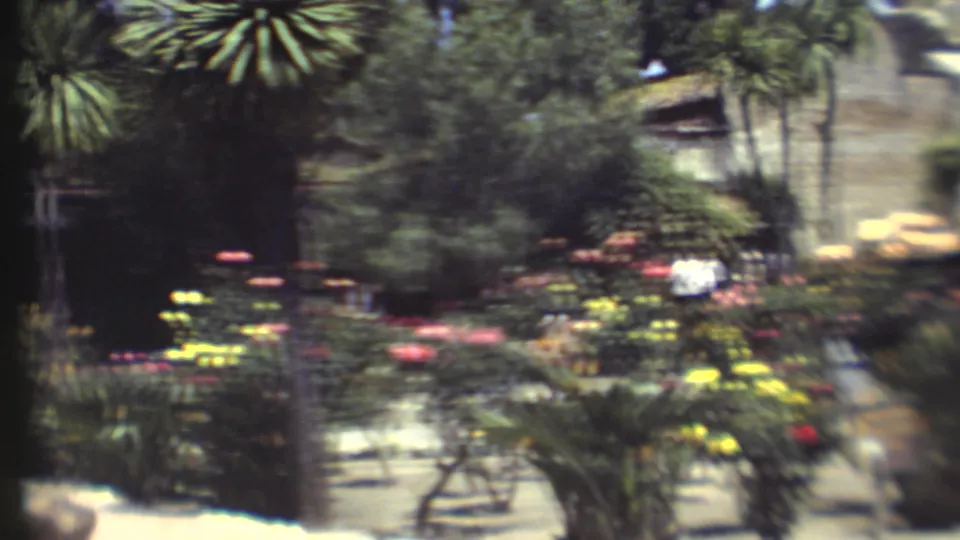 Soft 1972 Grand Canyon garden frame showing bright flowers, palm-like trees, shaded paths, and stone architecture in the background.