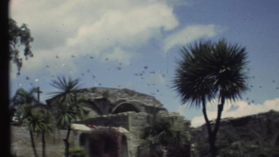 1972 Grand Canyon-area home movie view of a stone building with arched openings, palm-like trees, clouds, and birds in the sky.
