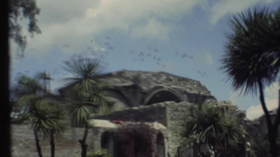 1972 Grand Canyon-area frame showing a stone building and wall, palm-like trees, soft sky, and scattered birds overhead.