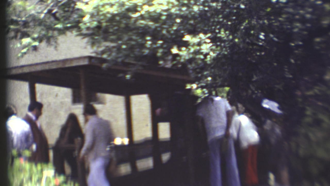People walk beside a shaded wooden structure amid trees and bright foliage.