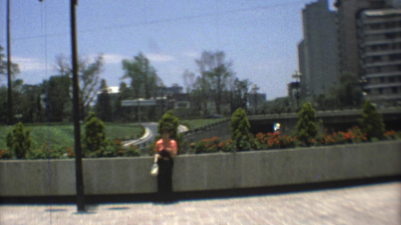 Woman in a red top stands near a concrete planter beside a winding road and modern buildings.