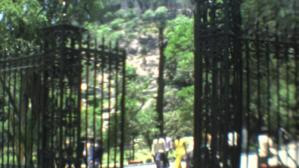 1973 Mexico City frame with ornate black iron gates, bright greenery, a rocky hillside, and pedestrians moving through the entrance.