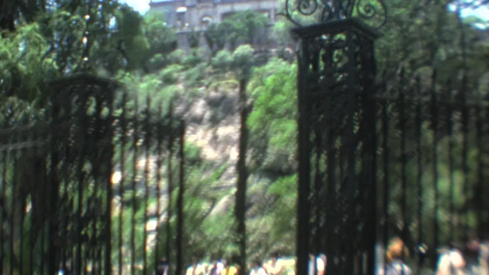 Soft 8mm view of tall iron gateposts and fencing, with leafy trees, hillside rock, and distant pedestrians near the gate.