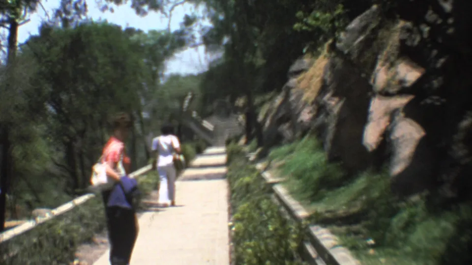 A home-movie view shows adults on a Mexico City park walkway with hedges, trees, low stone borders, and rocky landscaping.