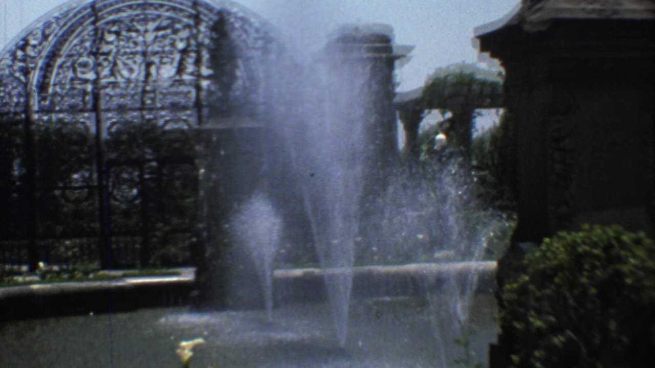 Fountain sprays in front of an ornate arched cemetery gate and stone pillars.