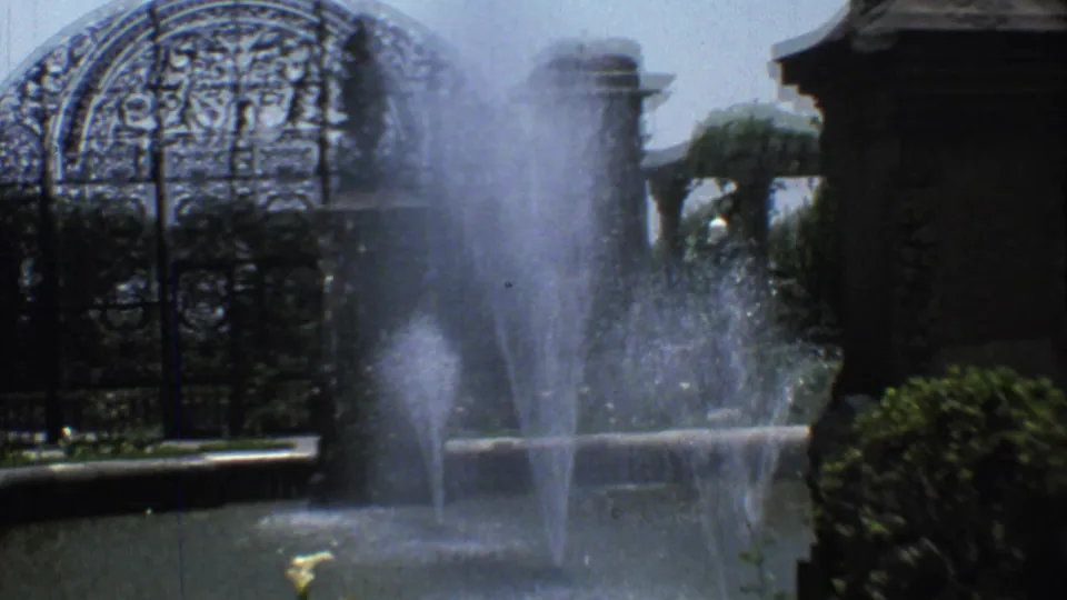 1973 Mexico City 8mm view of fountain jets rising before a decorative wrought-iron cemetery gate and stone entrance pillars.