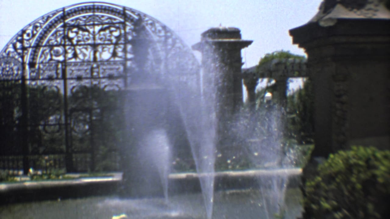 Fountain spray partially obscures a wrought-iron cemetery gate and stone entrance.