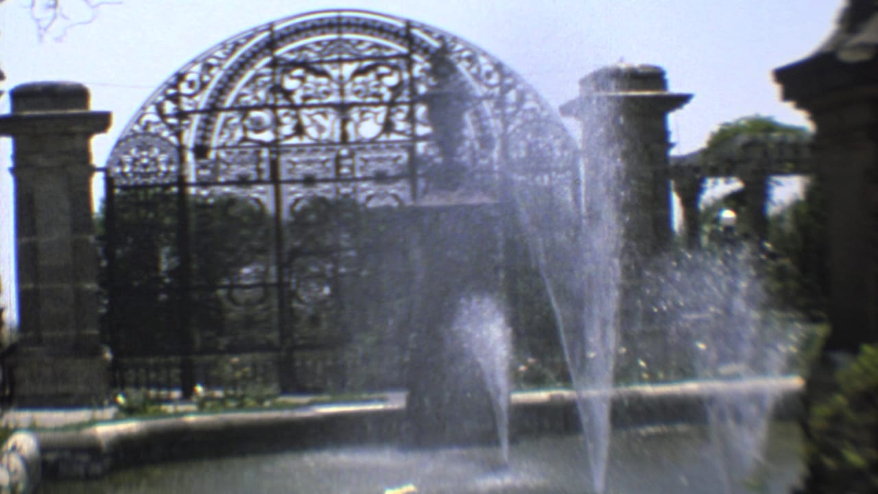 1973 8mm footage shows fountain water before a gothic-style cemetery gate in Mexico City, with a small distant visitor near the entrance.