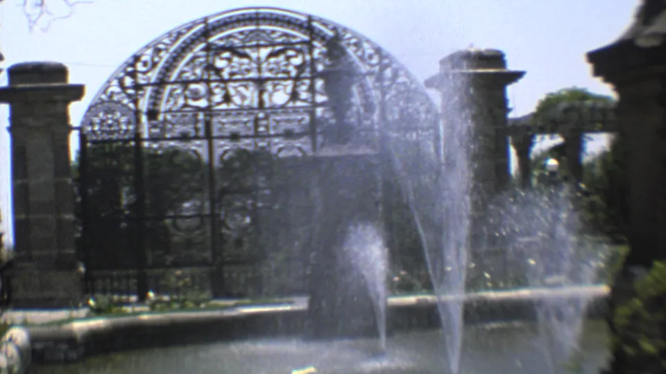 1973 8mm footage shows fountain water before a gothic-style cemetery gate in Mexico City, with a small distant visitor near the entrance.