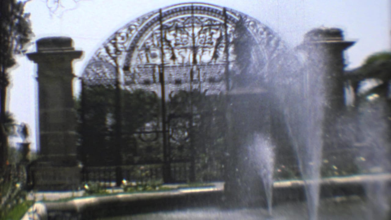 Final 1973 Mexico City frame holds on fountain spray, arched wrought-iron cemetery gate, stone entrance posts, and surrounding shrubs.