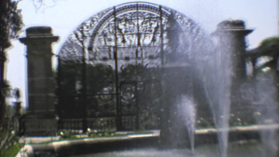 Final 1973 Mexico City frame holds on fountain spray, arched wrought-iron cemetery gate, stone entrance posts, and surrounding shrubs.