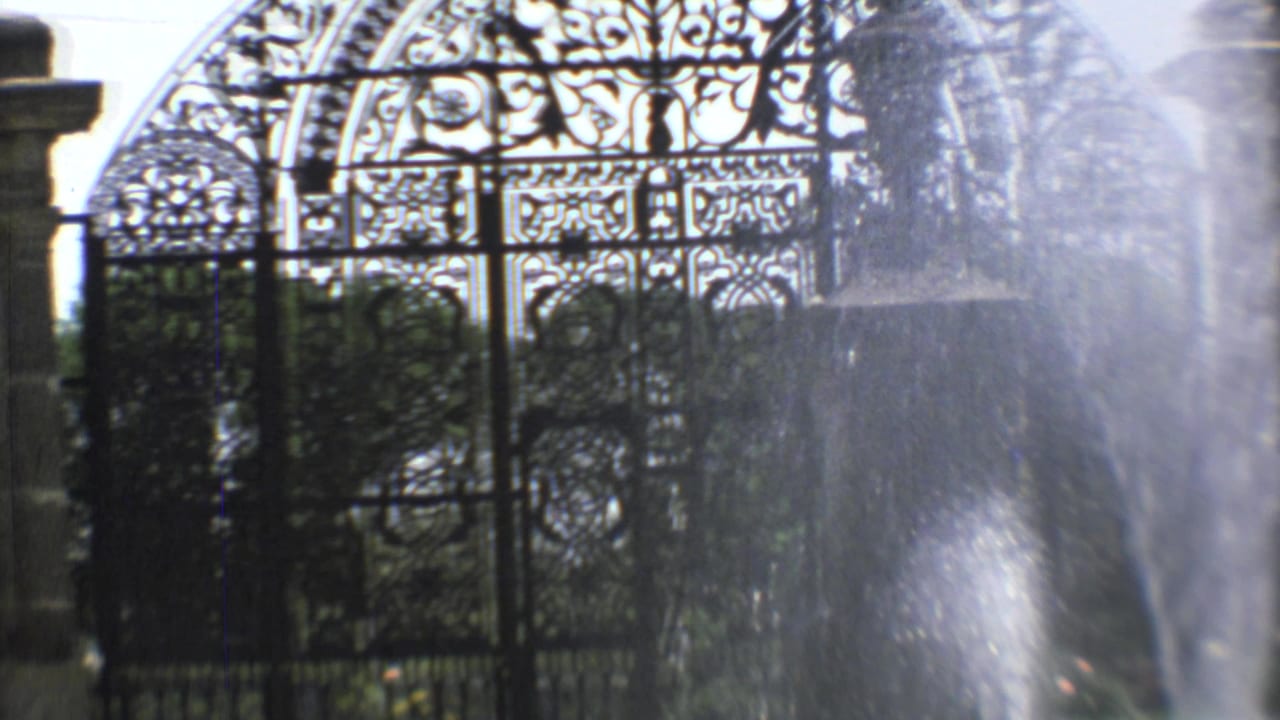 Ornate iron gate seen behind fountain spray and garden greenery.