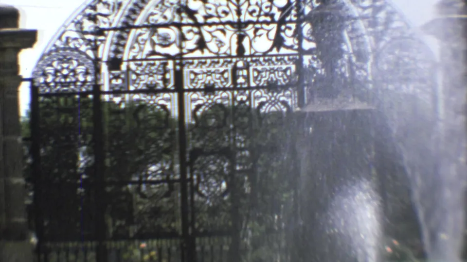 1973 Mexico City 8mm frame showing decorative iron gates partly obscured by fountain mist and shrubs.