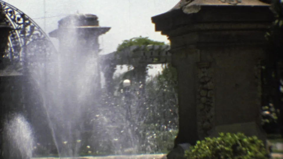 Mexico City garden entrance detail with stone pillar, greenery, and fountain spray on grainy 8mm film.