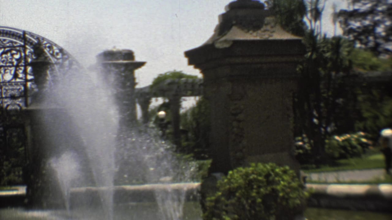 Fountain spray rises beside ornate stone pillars and wrought-iron gatework in a green garden.
