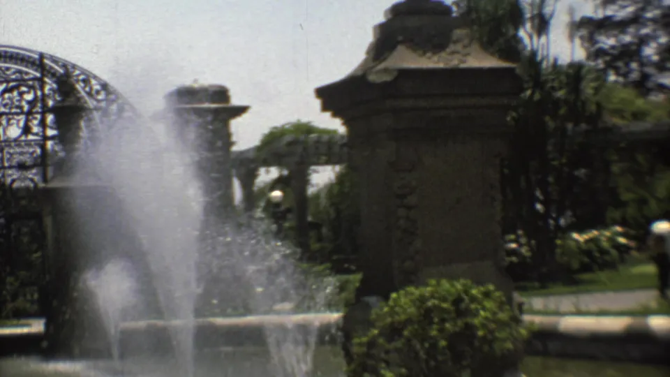 1973 Mexico City 8mm frame showing fountain spray, stone pillars, wrought-iron gatework, hedges, and sunlit garden plantings.