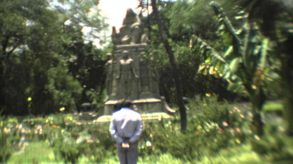 The frame shows a man from behind in a Mexico City garden, facing a tall stone statue partly framed by trees and flowering plants.